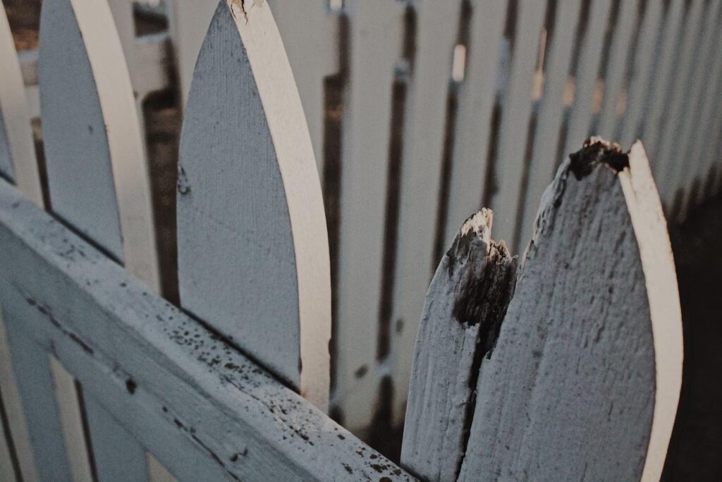 A close-up of a white wooden picket fence with one damaged and weathered plank, showing signs of decay and splintering.