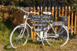 A white-painted bicycle used as a decorative signpost with handwritten signs for a bistro, bar, and rooftop terrace, placed beside a picket fence.