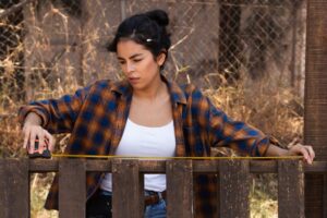 A woman in a plaid shirt uses a measuring tape to measure a wooden fence outdoors.