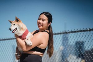 A smiling woman in athletic wear holds a dog in front of a chain-link fence.